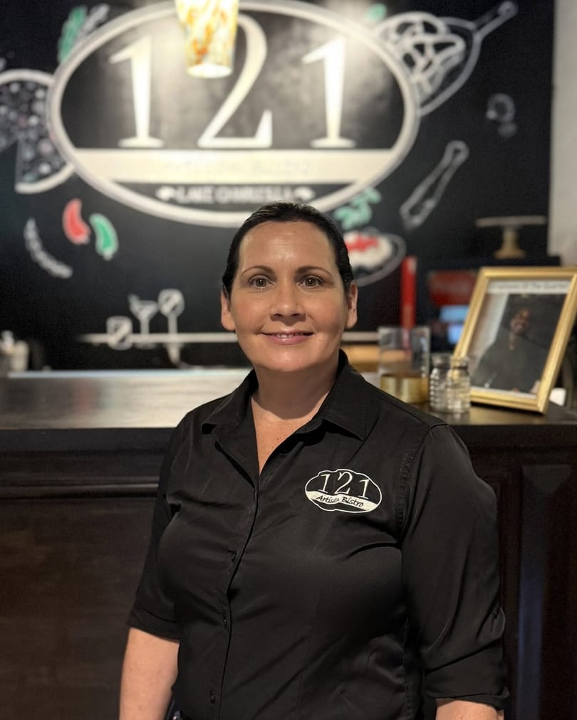 Smiling woman in a 121 Artisan Bistro uniform stands in front of a chalk art wall.