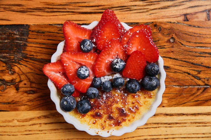 Creme brulee topped with sliced strawberries, blueberries, and powdered sugar in a white dish.