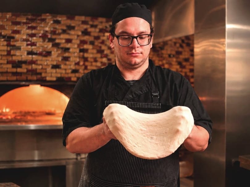 Chef in black uniform and glasses stretching pizza dough by a hot brick oven.