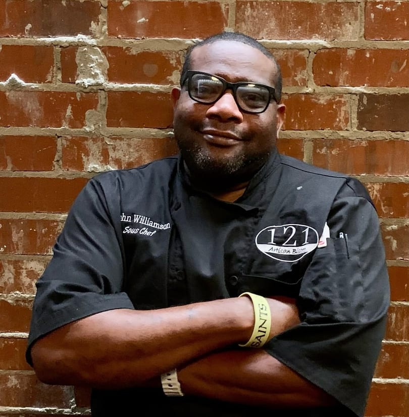 Smiling Sous Chef John Williamson in black chef coat with 121 Artisan Bistro logo, arms crossed, brick wall.