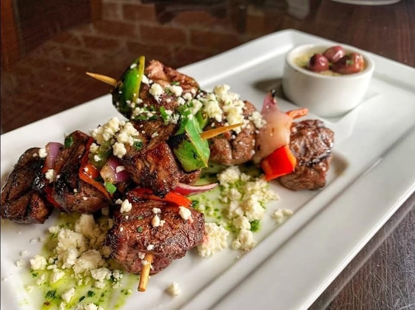 Two grilled meat and vegetable skewers with feta cheese, green sauce, and a side of olives.