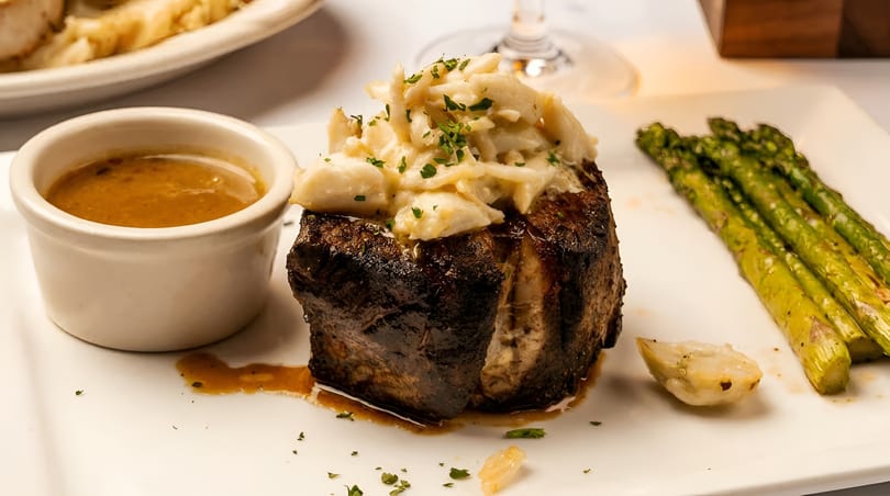 Seared steak topped with crab meat, served with a side of sauce and grilled asparagus.