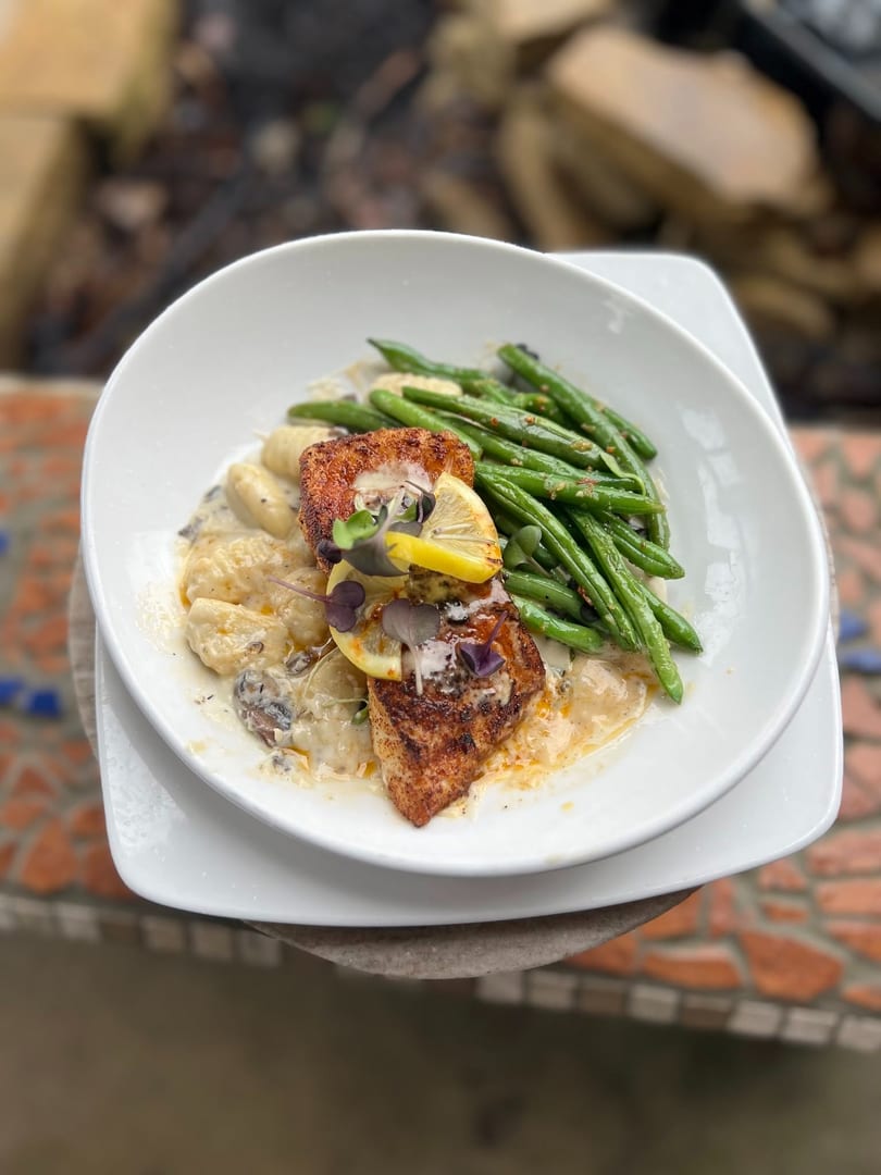 Seared fish, creamy gnocchi, and green beans with lemon garnish in a white bowl.