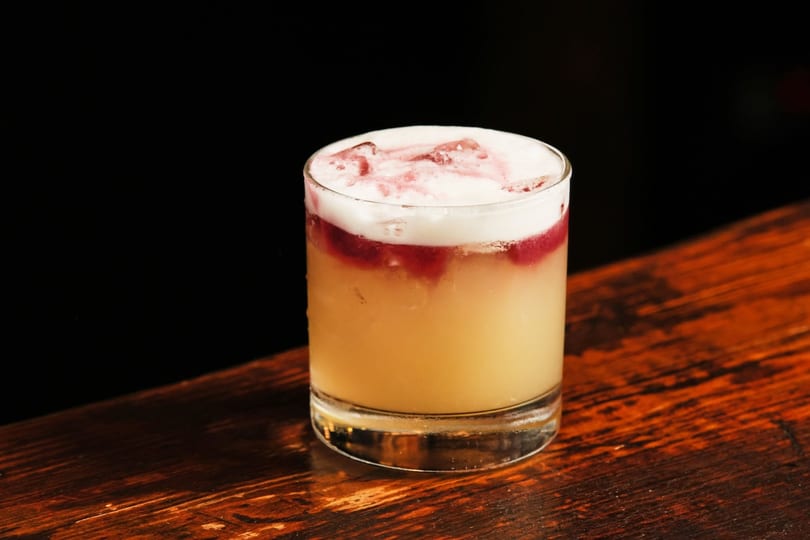 A frothy cocktail with red swirls sits on a dark wooden bar.