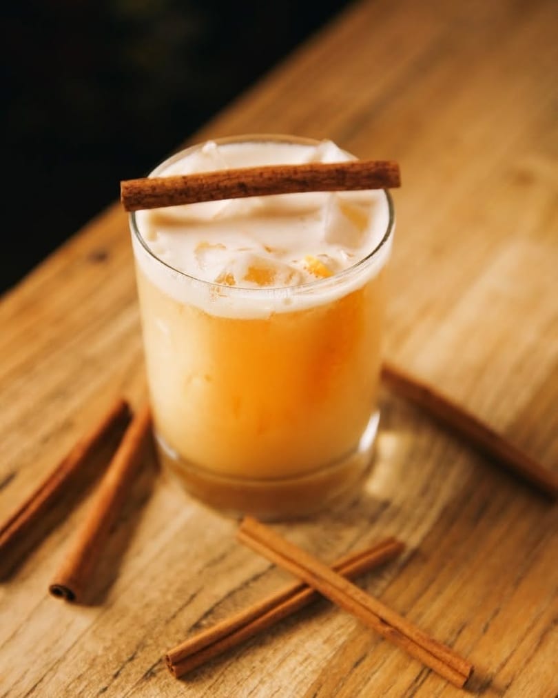 Iced cocktail with foam and a cinnamon stick on a wooden table with scattered cinnamon.