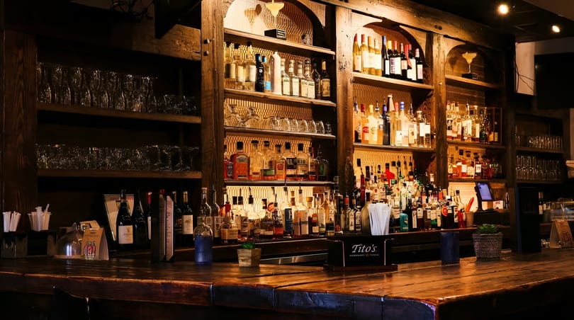 Warmly lit wooden bar counter with shelves full of liquor bottles and glassware.
