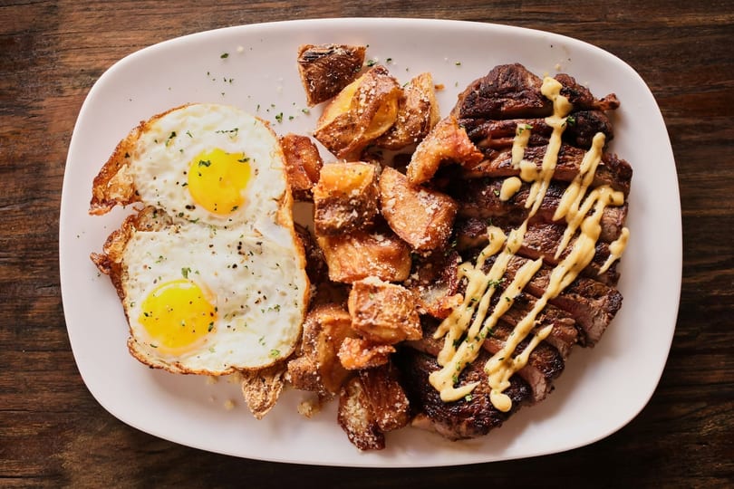 Sliced steak with creamy sauce, sunny-side up eggs, and parmesan roasted potatoes on a white plate.