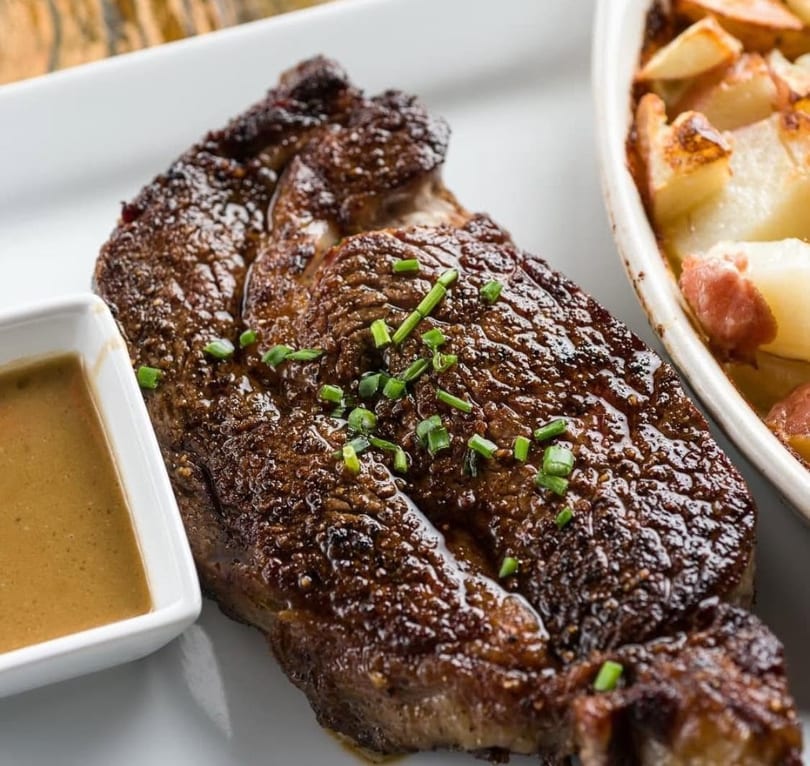 Grilled steak topped with chives, served with a side of sauce and roasted potatoes.