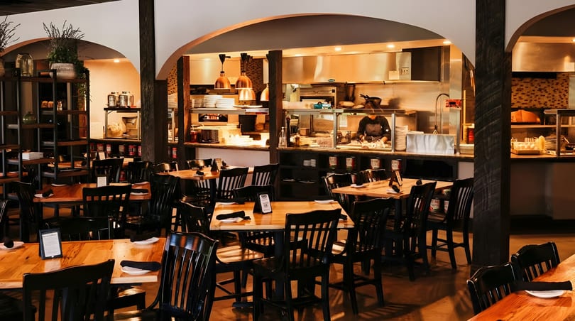 Cozy restaurant interior with wooden tables, black chairs, and an open kitchen where a chef works.
